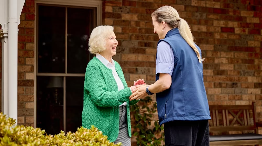 Support worker greeting an elderly woman outside her home, both smiling warmly while holding hands near a brick house facade.