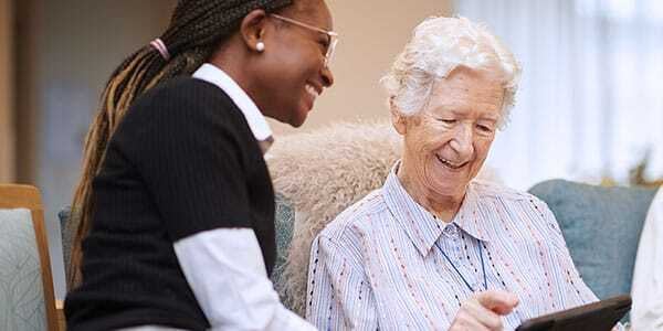 A smiling aged care worker assisting an elderly woman using a digital tablet, both seated and engaged in a friendly, supportive interaction.
