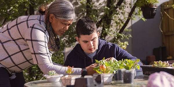 An older woman and a young man with disability engaging in a gardening activity together at an outdoor table, surrounded by potted plants.