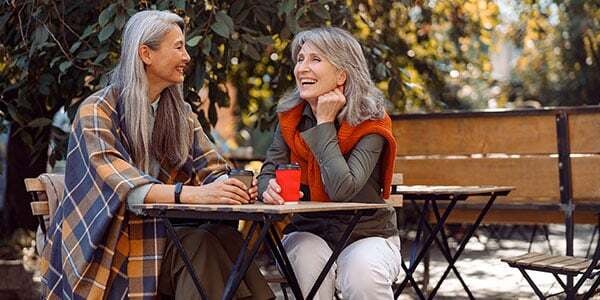 Two older women enjoying a warm drink and conversation together at an outdoor café, smiling and relaxed in a leafy, sunlit setting. Ask ChatGPT