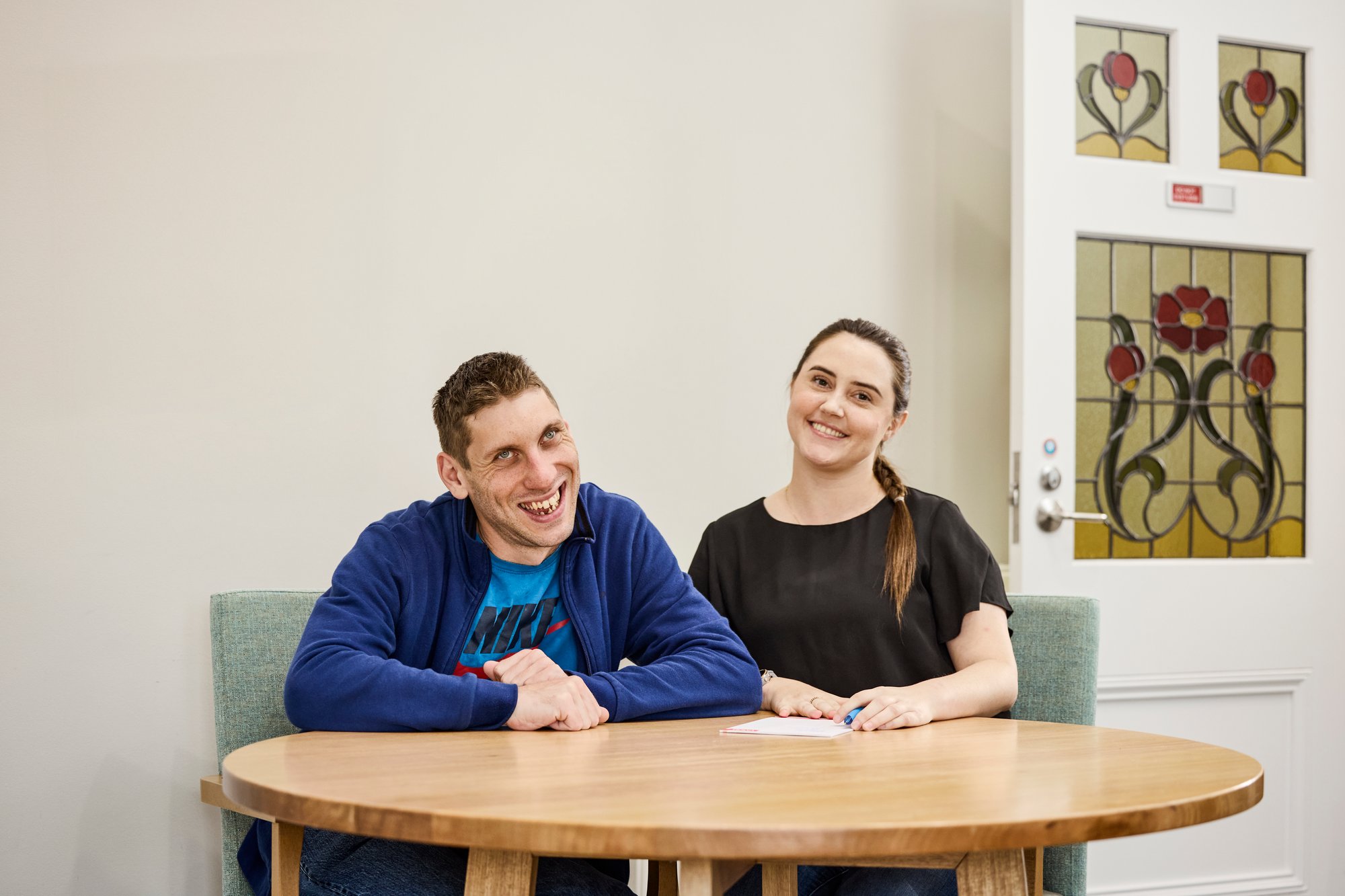 A man with a disability smiling, sitting at a table with a female support worker