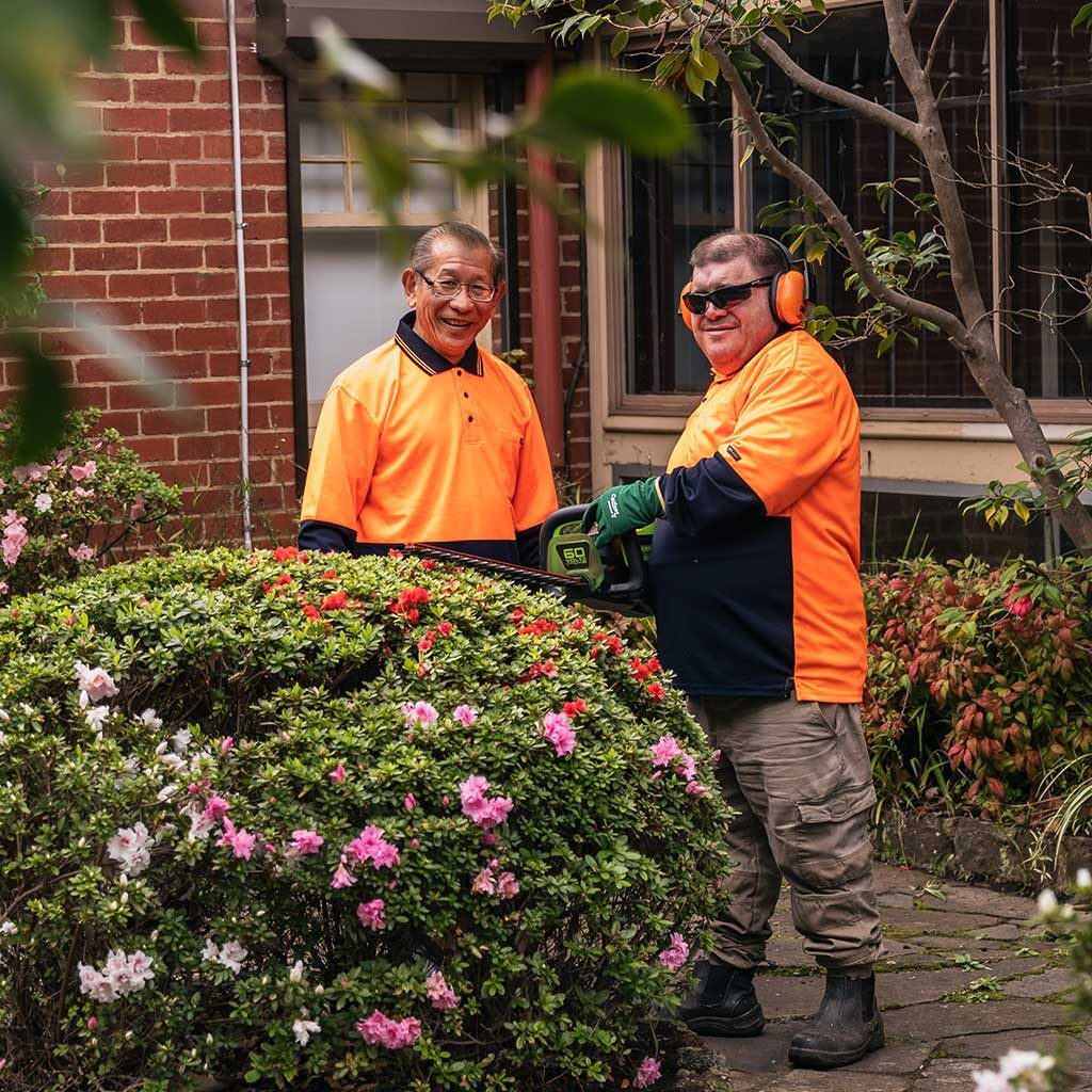 Two men in high-vis orange shirts smiling while trimming a flowering hedge in a garden outside a brick building.