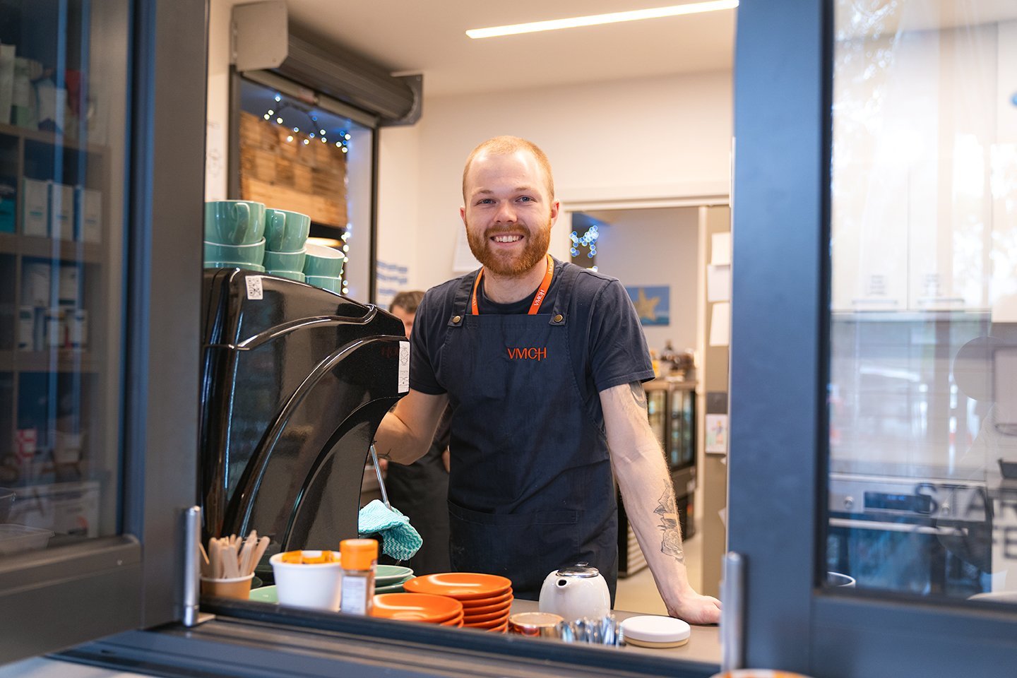 Barista smiling through a takeaway window while preparing coffee at a community café, wearing a VMCH apron and name badge.