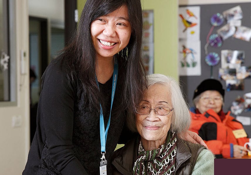 A smiling aged care worker with a name badge standing beside an elderly woman with glasses, both enjoying a moment together in a community room decorated with colourful artwork. Ask ChatGPT