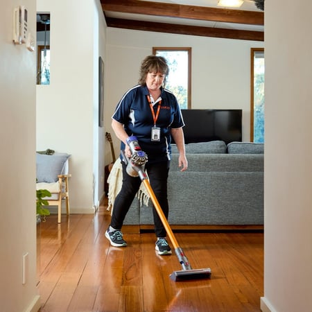A woman using a stick vacuum to clean a hardwood floor as part of an everyday service.