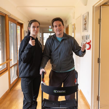 A man and a woman stand in a corridor and give a thumbs up to the camera
