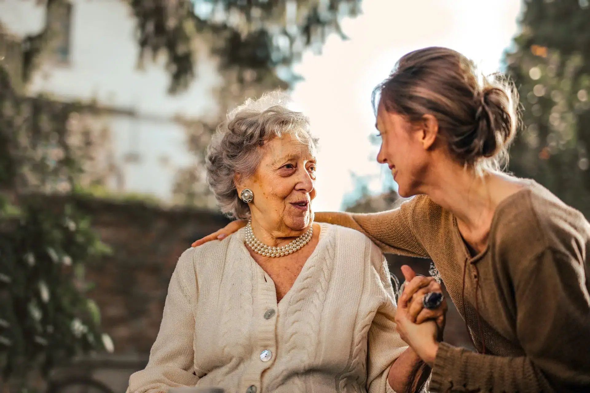An elderly woman sitting outdoors, smiling warmly while holding hands with a younger woman who leans in with a caring gesture, highlighting a moment of connection and support.