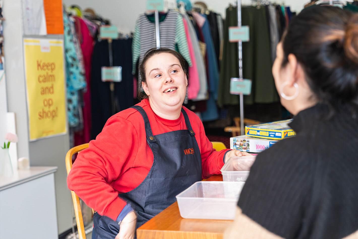 Young woman wearing a red shirt and VMCH apron smiles while chatting with a customer inside an op shop, with second-hand clothes and puzzles in the background.