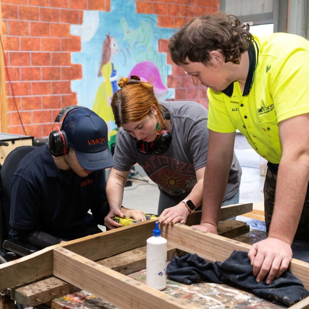 Three people collaborating on a woodworking project in a workshop, with one person in a wheelchair using a power tool under supervision. A colourful mural is painted on the wall behind them.