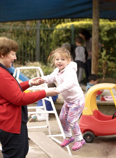 A young girl jumping and holding hands with a caregiver in an outdoor early learning centre playground, surrounded by play equipment and other children.