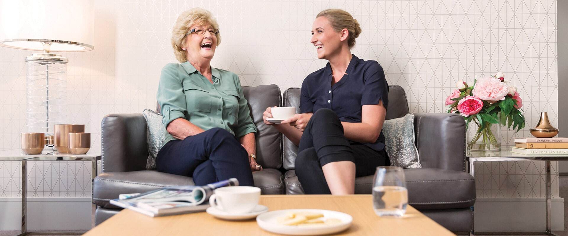 An older woman and a younger woman sitting together on a couch, smiling and chatting over cups of tea in a bright, modern living room with flowers and decor, creating a warm and welcoming atmosphere.