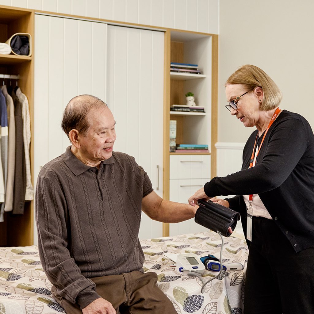 An older man sitting in his bedroom while a nurse takes his vitals as part of the EOL pathway.