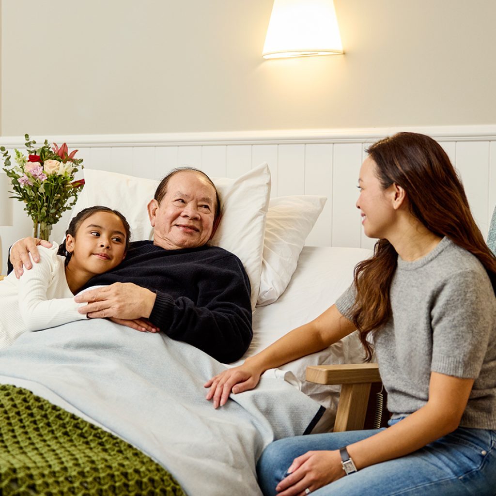 An older man in bed with a woman sitting next to him and a child leaning over giving him a hug.