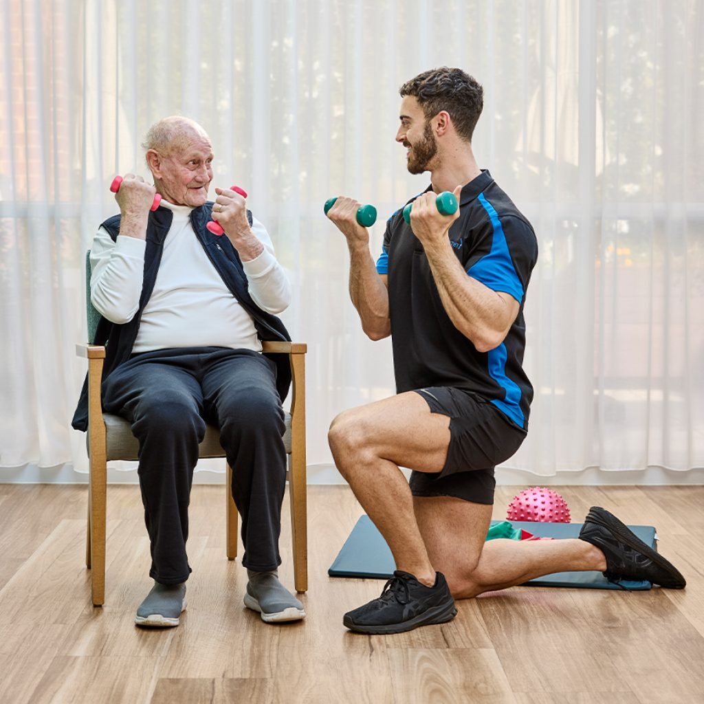 A younger man running a physiotherapist session with an older man, both are holding weights.