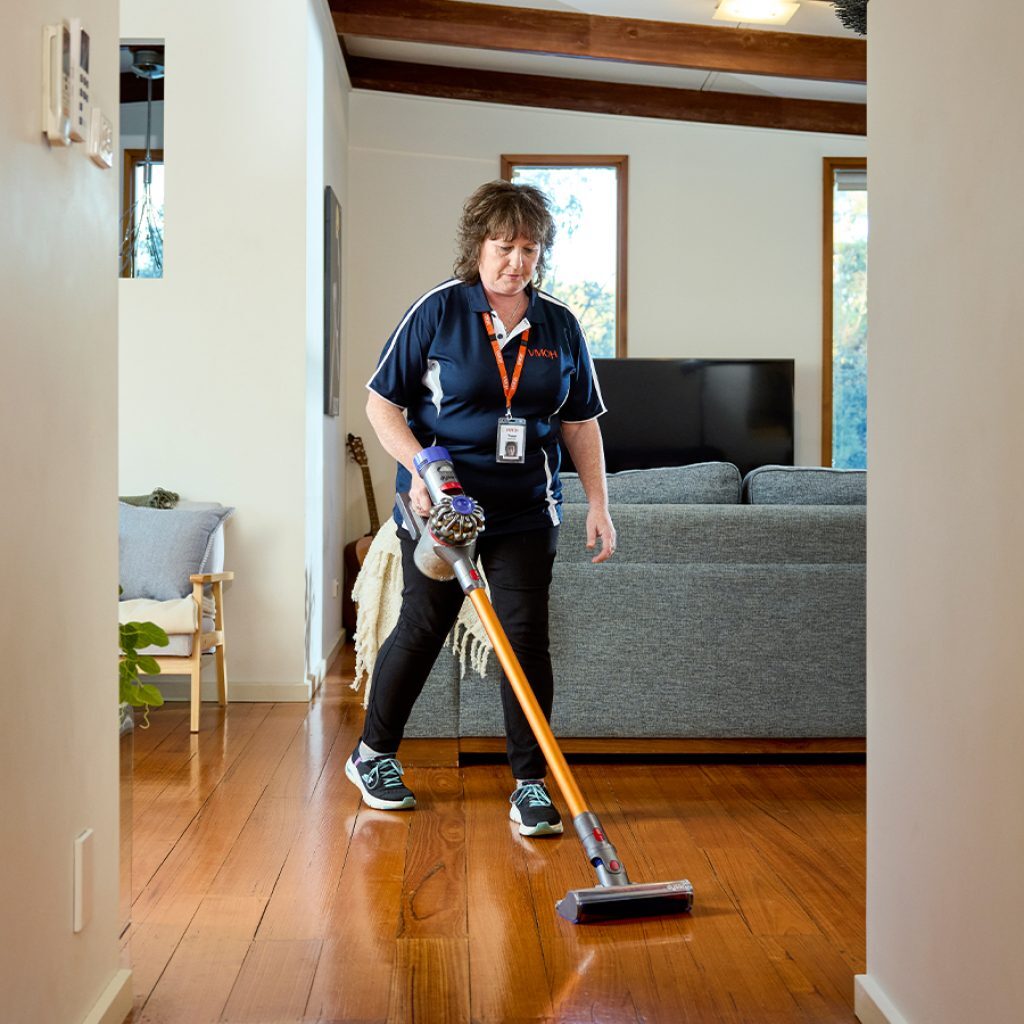 A woman using a stick vacuum to clean a hardwood floor as part of an everyday service.