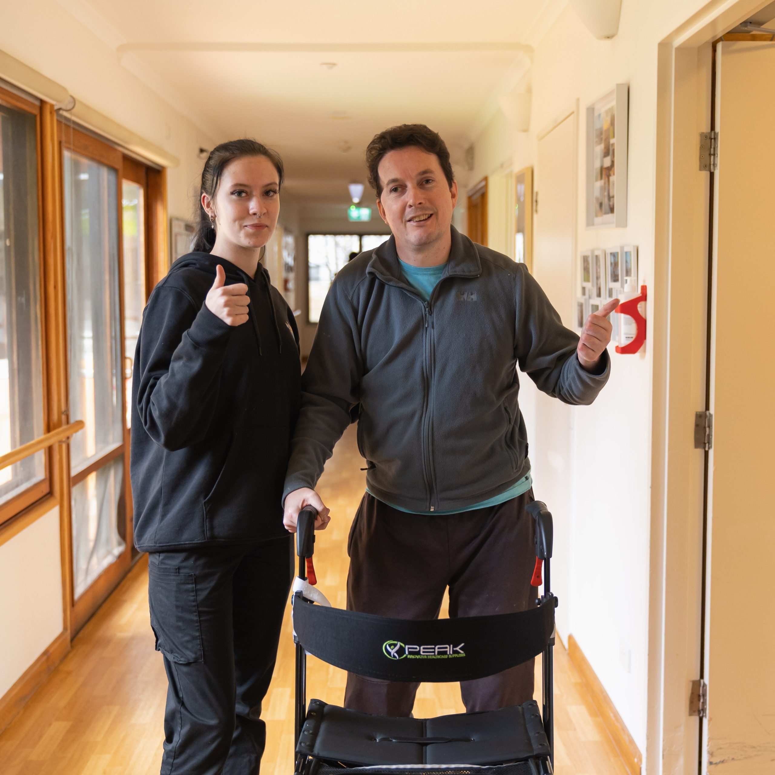 A man and a woman stand in a corridor and give a thumbs up to the camera