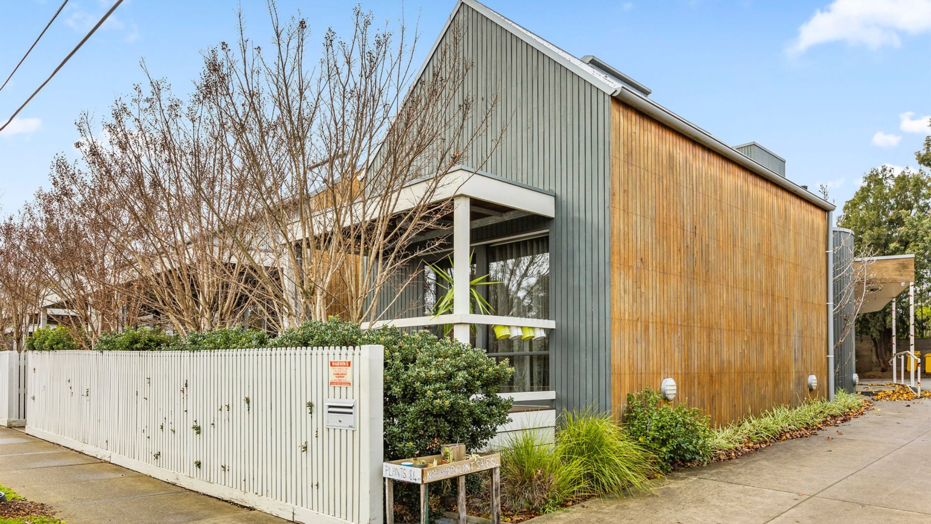 The front of a residence. The home is brown and grey in colour and there is a white picket fence in the foreground.